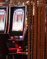 A pair of casino slot machines, each with detailed screens displaying various game outcomes and jackpot amounts. The chairs in front of the slot machines are dark and cushioned for comfort. The background features a hanging curtain made of golden ropes and other casino decorations are visible.