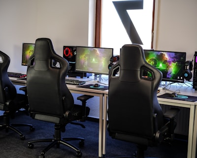A row of gaming stations equipped with large monitors, gaming chairs, keyboards, and headphones. The setup is organized and ready for gaming, featuring a colorful screen display. The room has a modern and tech-savvy ambiance.