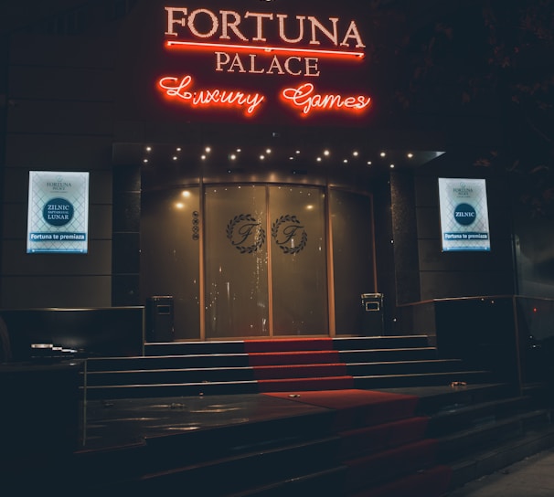 A neon-lit casino entrance with a red carpet leading up to large double doors. The sign reads 'Fortuna Palace Luxury Games' in bright red and white neon lights. The dimly lit atmosphere suggests a night-time setting. There are two digital displays on either side of the entrance showing promotional content.