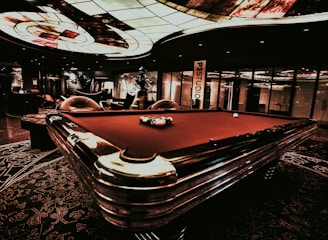 A luxurious interior features a billiard table with balls arranged on it. The ceiling has an intricate stained glass design, and the space is dimly lit, creating a sophisticated atmosphere. Comfortable chairs are placed around the area, with some decorative plants and a lamp adding to the elegance.