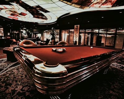A luxurious interior features a billiard table with balls arranged on it. The ceiling has an intricate stained glass design, and the space is dimly lit, creating a sophisticated atmosphere. Comfortable chairs are placed around the area, with some decorative plants and a lamp adding to the elegance.